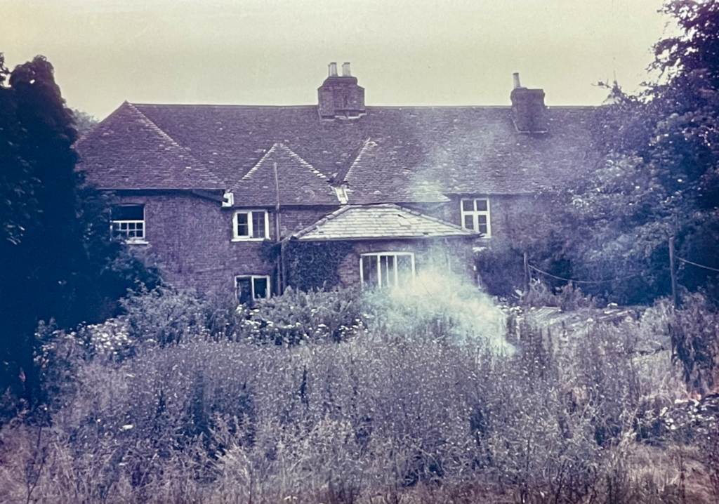 Sepia photo of brown Manor Farm building. In the foreground, tall grass is visible. There is smoke coming up amidst the weeds