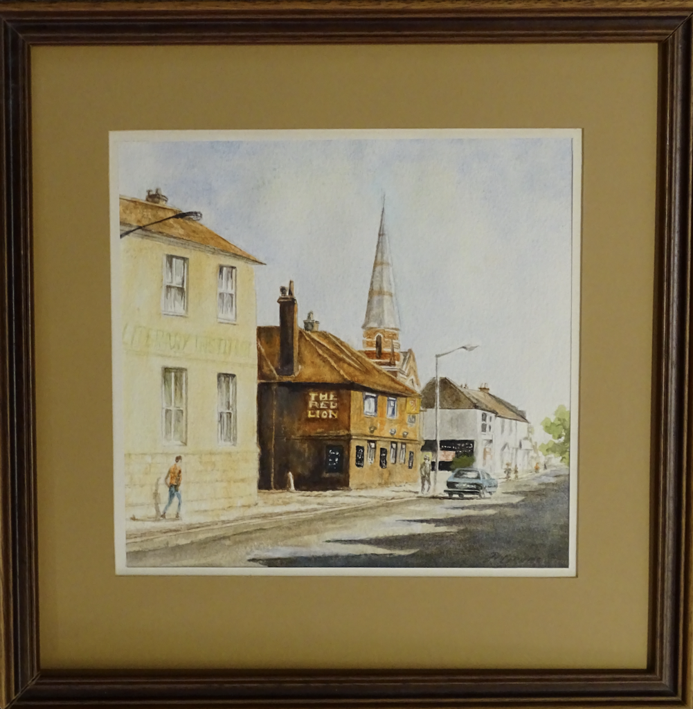 dark-wooden framed watercolour of Egham High Street. View of Literary Institute and Red Lion buildings. There is a figure in front of Lit Institute with orange shirt and blue jeans, and a figure in grey next to a street lamp and a blue car in front of red lion. Grey road visible from foreground to background on the left side of the painting. Shops, tree and church spire visible in the background