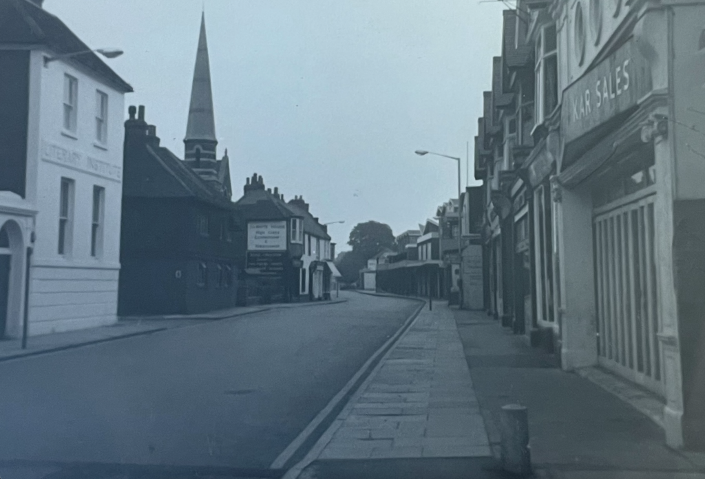 black and white photo of same high street view as previous image. This photo also shows shop windows on the right hand side